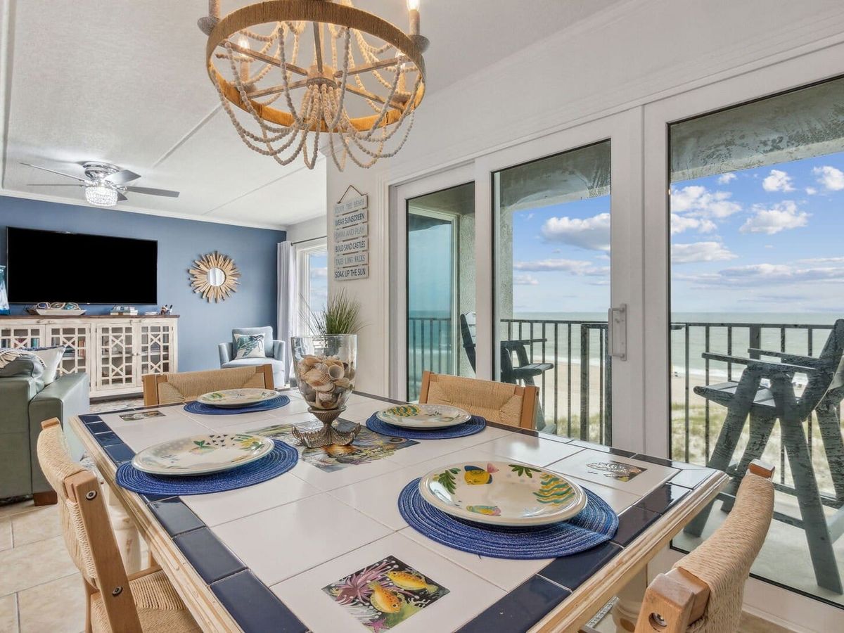 Bright beachfront condo with a tile-top dining table and tropical-themed plates, overlooking the ocean. Large glass doors open to a balcony with panoramic views of the sand and sea—perfect for breakfast with a view.