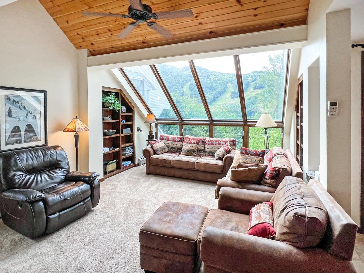 A cozy mountain-view living room with large angled windows showcasing a lush ski slope, featuring plush brown sofas, a leather recliner, built-in bookshelves, and a wood-paneled ceiling with a ceiling fan.