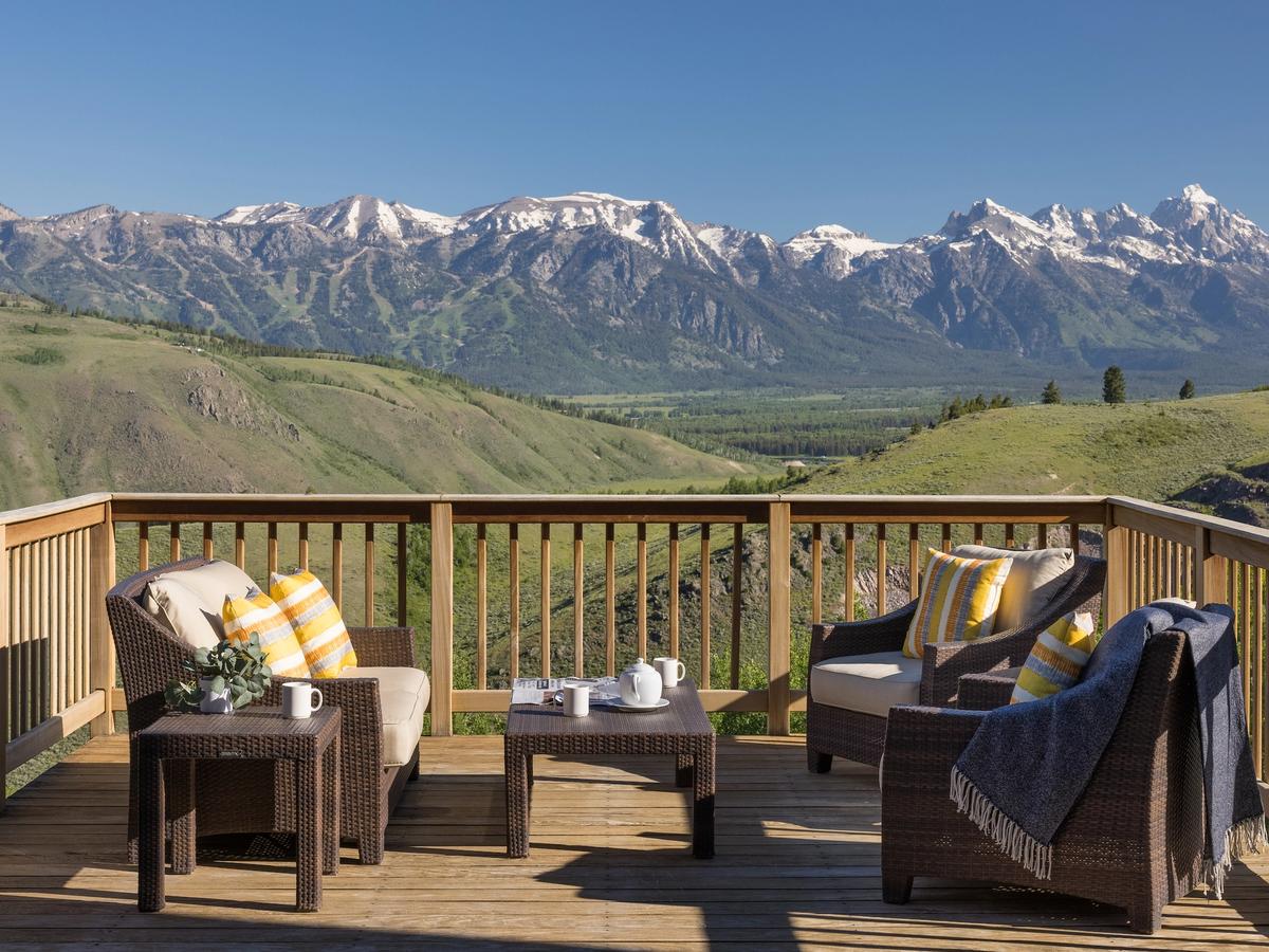 Cozy mountain-view deck with wicker patio furniture, yellow-striped pillows, and a coffee setup overlooking a green valley and snow-capped peaks.