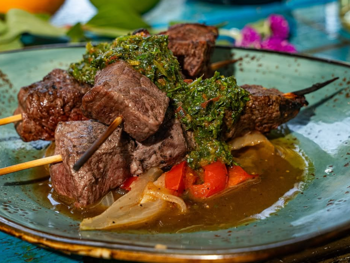 Skewers of grilled beef served over roasted peppers and onions, topped with a generous portion of chimichurri sauce on a green ceramic plate.