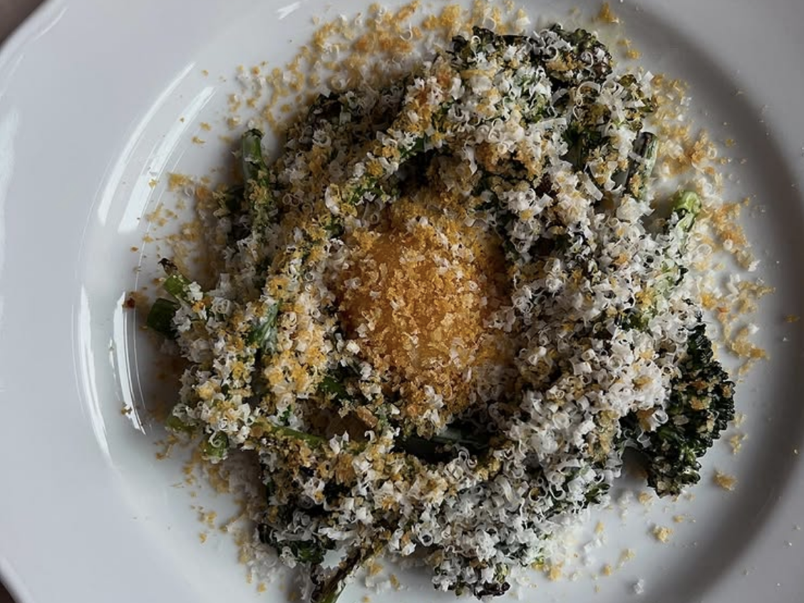 A white plate featuring charred broccolini generously topped with finely grated cheese and golden breadcrumbs, artfully arranged in a ring around a bright yellow egg yolk center.