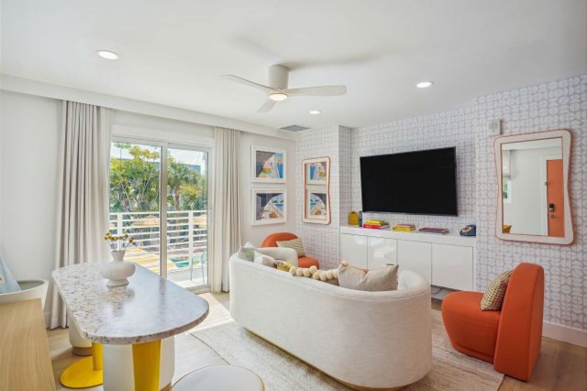 Modern and playful living space featuring a curved white sofa, bold orange accent chairs, and geometric wallpaper with a mounted flat-screen TV.