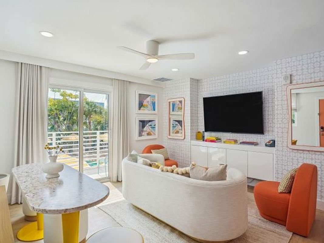 Modern and playful living space featuring a curved white sofa, bold orange accent chairs, and geometric wallpaper with a mounted flat-screen TV.