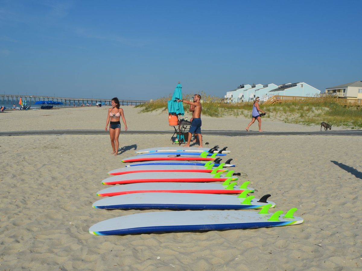 Surfboards are lined up on the sand at Carolina School of Surf, ready for a beach lesson. The bright boards and ocean setting make it a fun spot for beginners to learn.