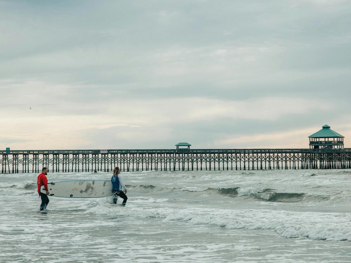 Two people carry a surfboard through the waves near a long wooden pier stretching into the ocean under a cloudy sky.