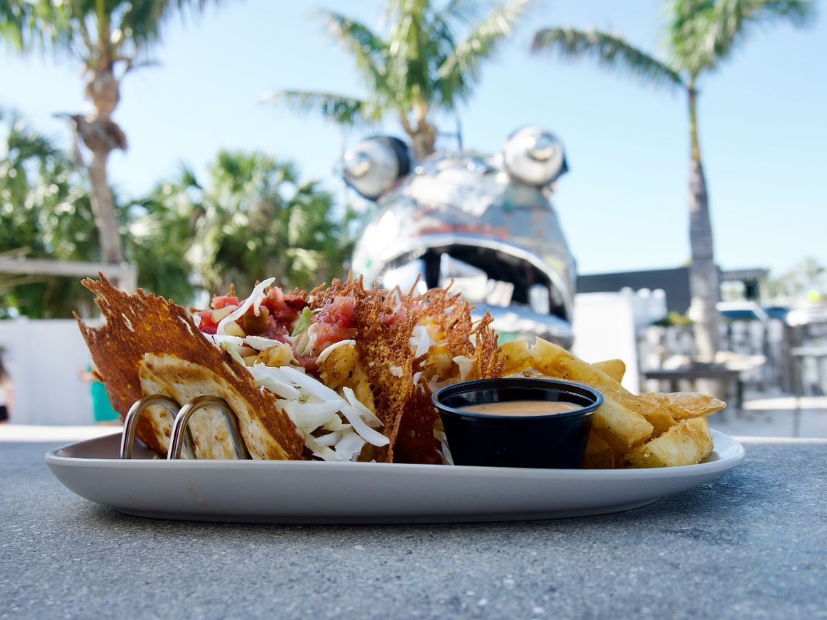 Fish tacos served under sunny skies with crispy shells, shredded cabbage, and salsa beside golden fries. The playful outdoor setup makes every meal feel like a beach party.