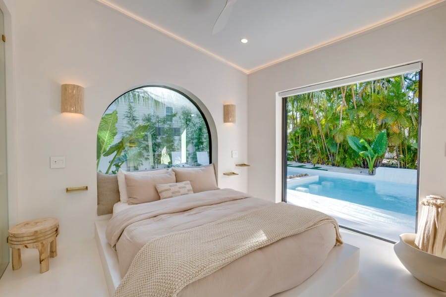 A bright, airy bedroom with soft neutral bedding and a wide window overlooking a private pool surrounded by palm trees. The minimalist design blends luxury and calm, perfect for a tropical escape.