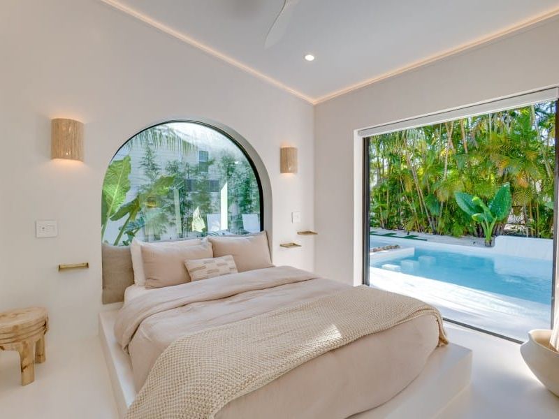 A bright, airy bedroom with soft neutral bedding and a wide window overlooking a private pool surrounded by palm trees. The minimalist design blends luxury and calm, perfect for a tropical escape.