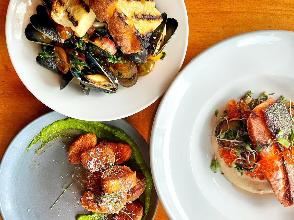 An overhead spread of three elegantly plated dishes rests on a warm wood table, featuring mussels in broth with grilled bread, crispy gnocchi over vibrant green sauce, and seared salmon with roe and microgreens. The composition highlights the restaurant's refined, ingredient-driven cooking.
