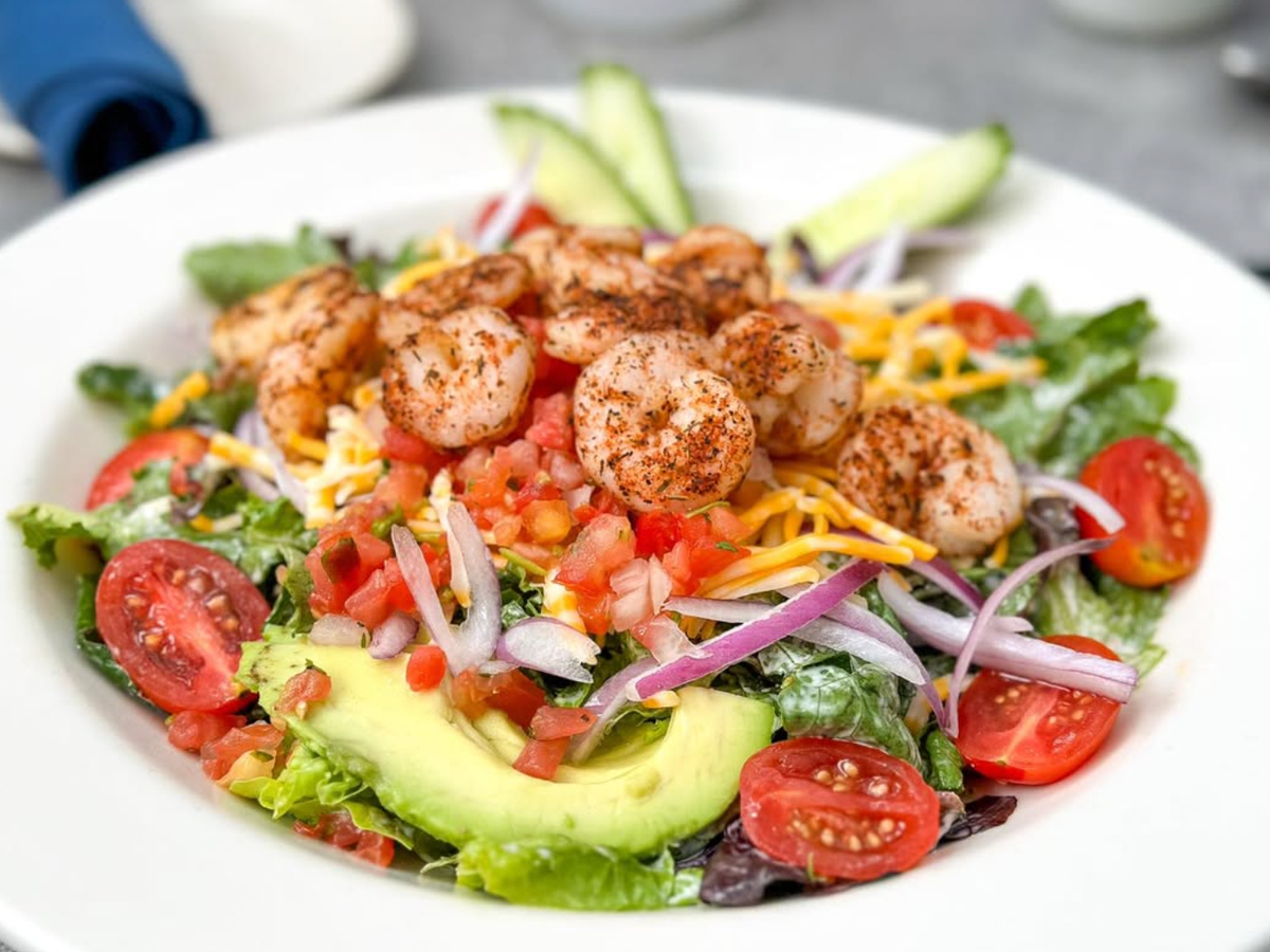 A fresh salad topped with seasoned shrimp, sliced avocado, cherry tomatoes, shredded cheese, red onion, and pico de gallo, served on a bed of mixed greens with cucumber slices.