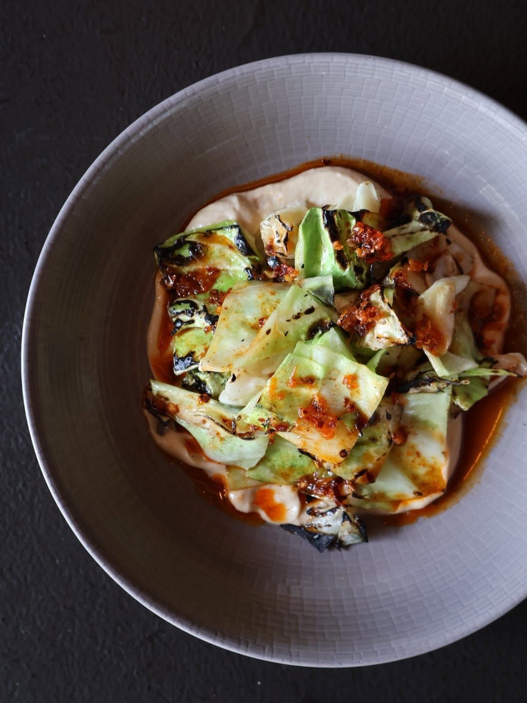Charred wedges of grilled cabbage glisten with spiced chili oil over a creamy pool of whipped sauce in a pale ceramic bowl. The moody lighting and minimalist plating highlight the dish's smoky, elegant presentation.