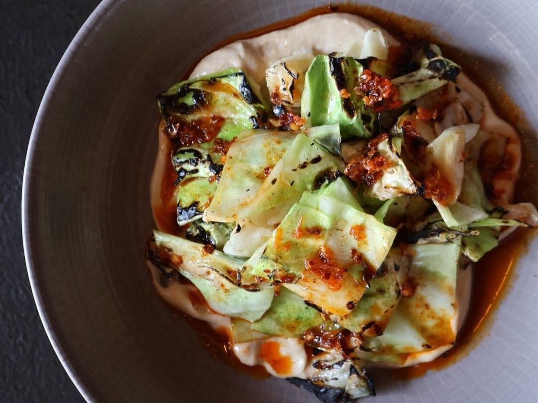 Charred wedges of grilled cabbage glisten with spiced chili oil over a creamy pool of whipped sauce in a pale ceramic bowl. The moody lighting and minimalist plating highlight the dish's smoky, elegant presentation.