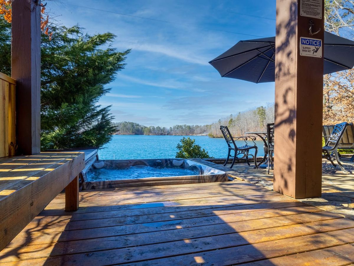 The Hangout at Lake Keowee is a lakefront escape with warm wood interiors, comfy furniture, and full glass doors looking out over the water. A private dock and hot tub make it the perfect retreat.