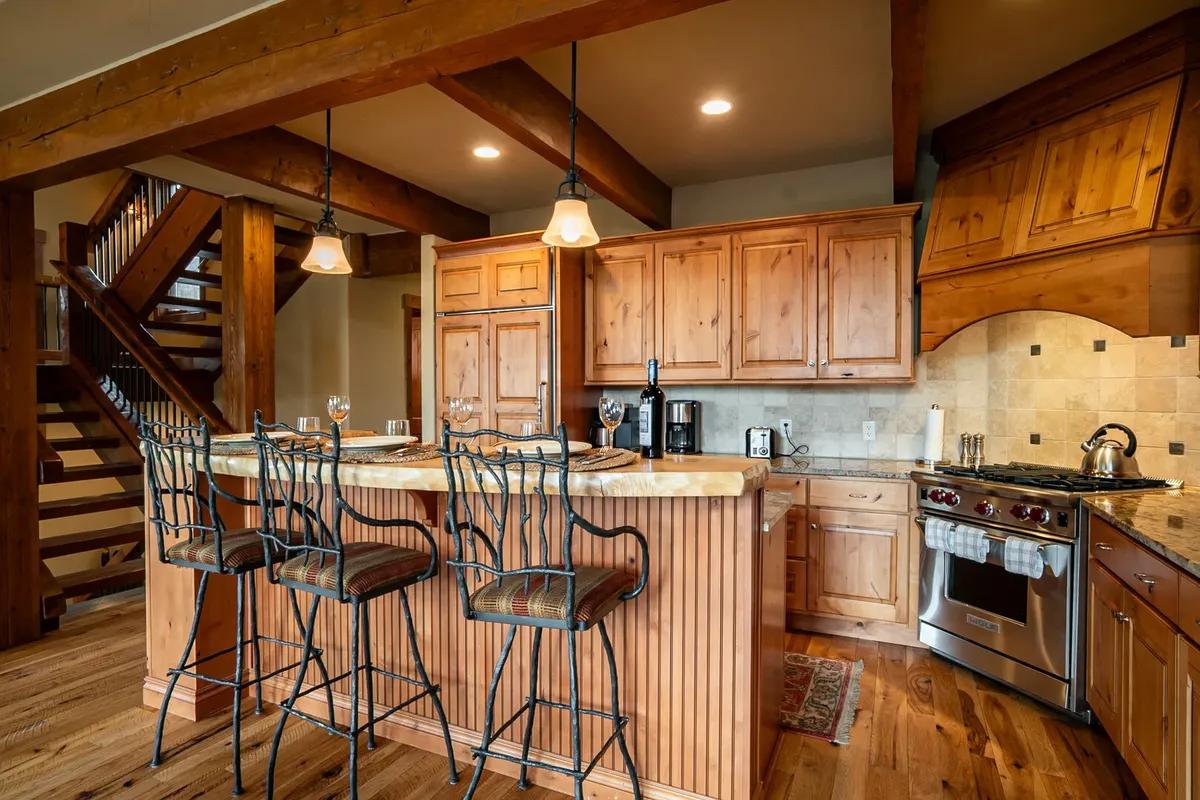 A cozy rustic kitchen with natural wood cabinetry, granite countertops, stainless steel appliances, a central island with barstool seating, and exposed wooden beams adding mountain lodge charm.