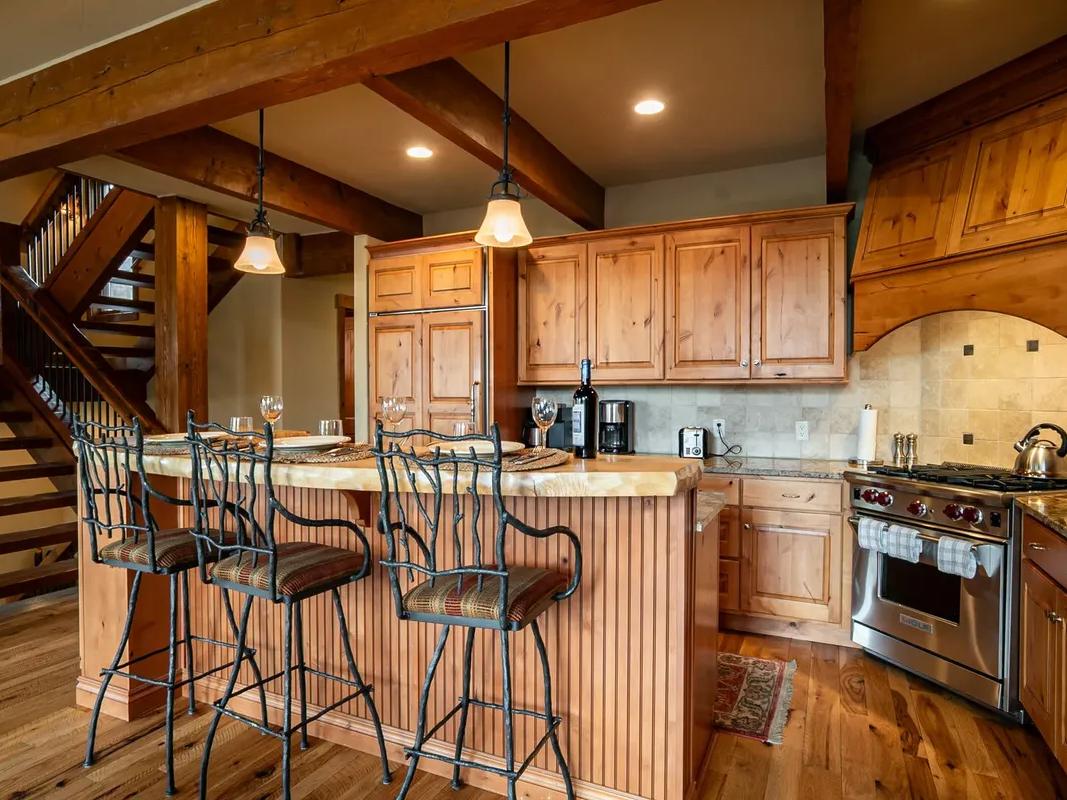 A cozy rustic kitchen with natural wood cabinetry, granite countertops, stainless steel appliances, a central island with barstool seating, and exposed wooden beams adding mountain lodge charm.