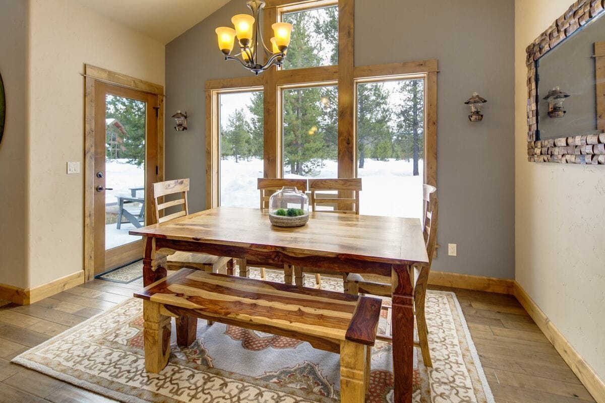 A cozy mountain retreat surrounded by tall pines, featuring an inviting firepit with outdoor seating overlooking a peaceful river. The spacious deck offers plush lounge seating and dining space perfect for barbecues or watching sunsets. Inside, warm wood finishes and high ceilings create a comfortable, lodge-style living space ideal for families or groups.