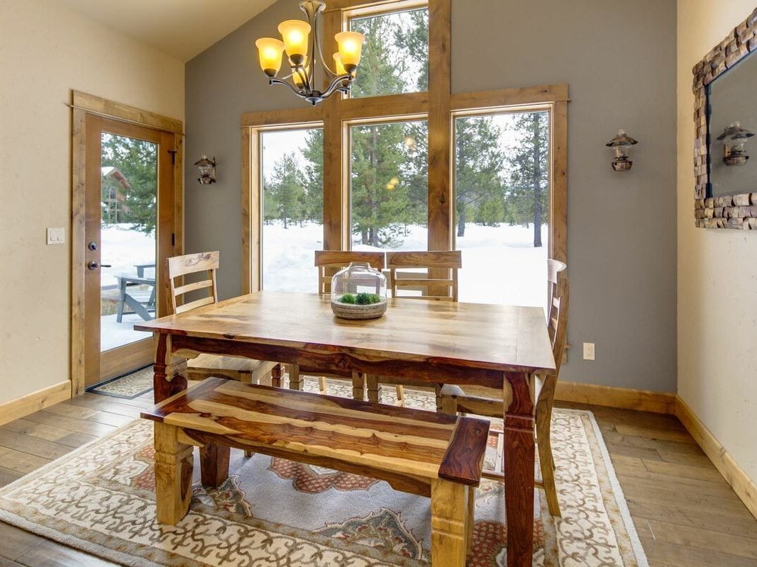 A cozy mountain retreat surrounded by tall pines, featuring an inviting firepit with outdoor seating overlooking a peaceful river. The spacious deck offers plush lounge seating and dining space perfect for barbecues or watching sunsets. Inside, warm wood finishes and high ceilings create a comfortable, lodge-style living space ideal for families or groups.