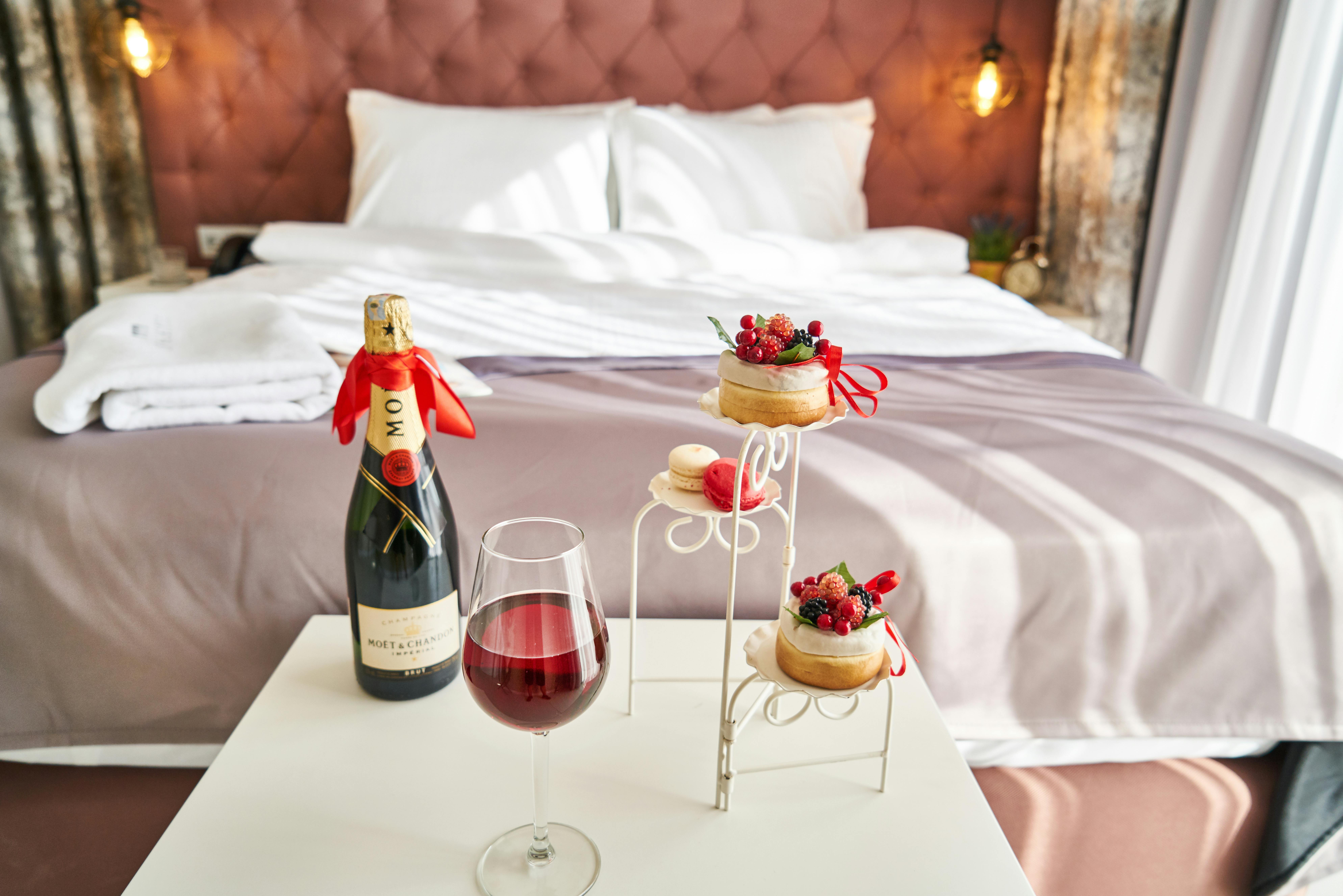 A bottle of champagne, desserts, and a glass of wine sit on a bedside table in a luxurious hotel room. The setup evokes indulgence and relaxation for a perfect morning or romantic evening.