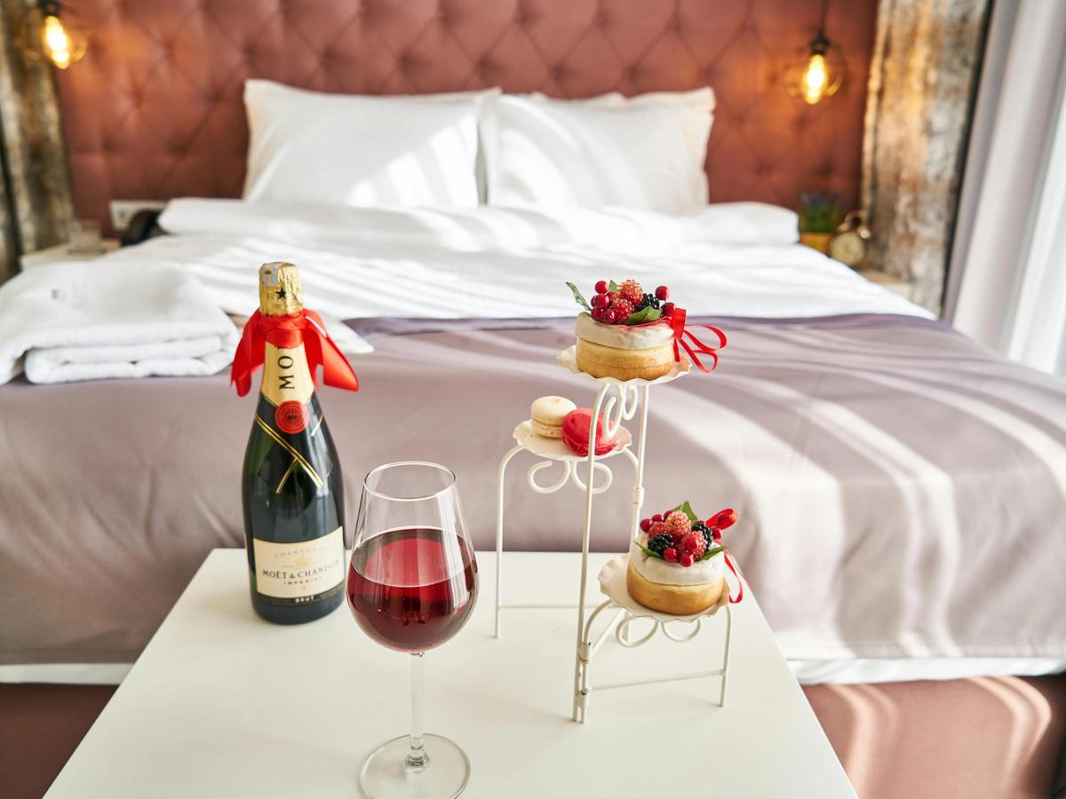 A bottle of champagne, desserts, and a glass of wine sit on a bedside table in a luxurious hotel room. The setup evokes indulgence and relaxation for a perfect morning or romantic evening.