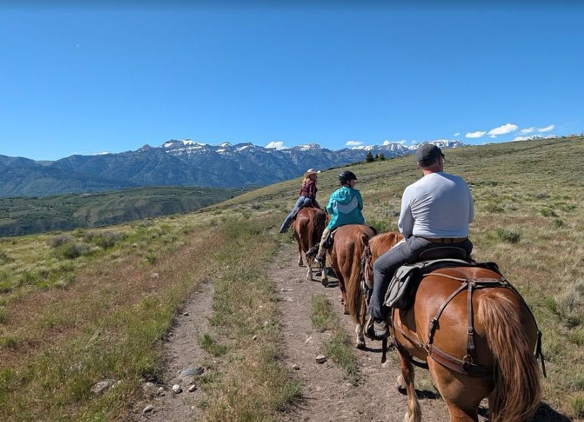 A group of riders on horseback follows a dirt trail across rolling green hillsides at Spring Creek Ranch, with the snow-dusted Teton Range rising in the distance. Clear blue skies stretch above the wide-open sagebrush landscape.