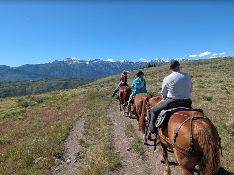 A group of riders on horseback follows a dirt trail across rolling green hillsides at Spring Creek Ranch, with the snow-dusted Teton Range rising in the distance. Clear blue skies stretch above the wide-open sagebrush landscape.