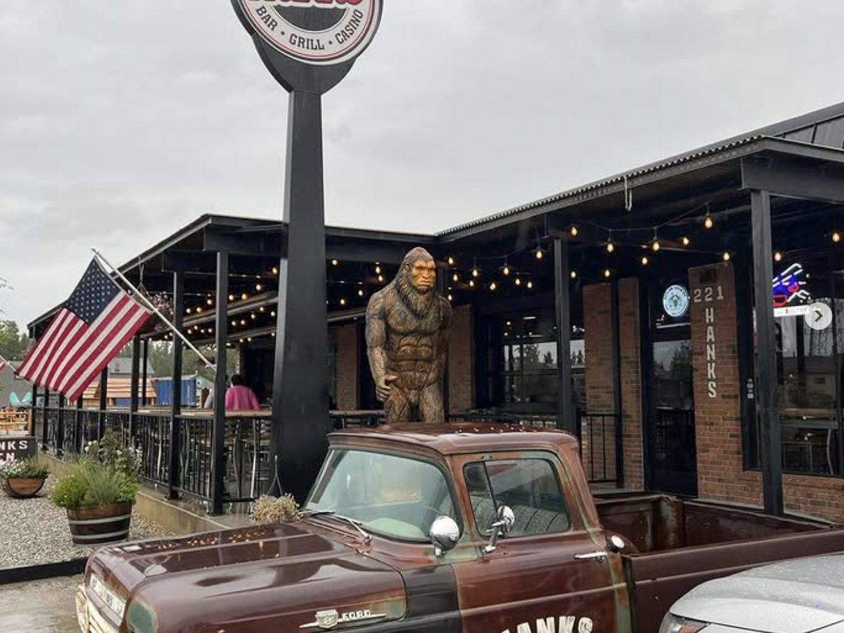 Exterior of Hank’s Bar & Grill featuring a vintage truck, an American flag, string lights, and a large Bigfoot statue near the entrance.