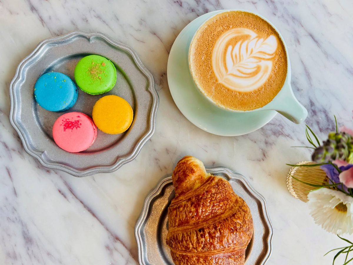 A latte with delicate rosetta art sits on a marble countertop beside a flaky golden croissant and a plate of pastel macarons in blue, green, pink, and yellow. A small bouquet of fresh flowers adds a soft, elegant touch to the bright café setting.