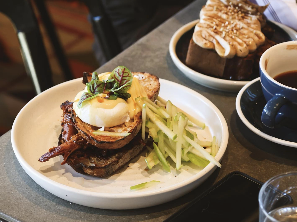 A beautifully plated brunch featuring a pulled pork eggs Benedict on toasted sourdough with a poached egg, hollandaise, and microgreens, served with julienned green apple; in the background is a cinnamon roll with frosting and a cup of black coffee.