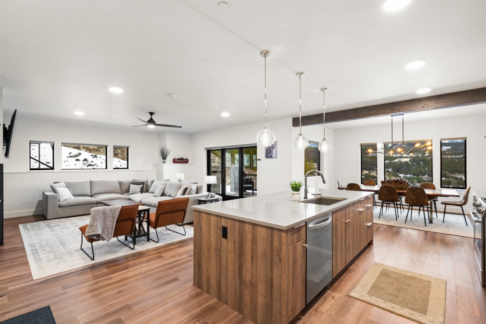 A modern open-concept kitchen and living area with a wood island, pendant lighting, and a spacious gray sectional opens to a dining space with large windows overlooking mountain views.