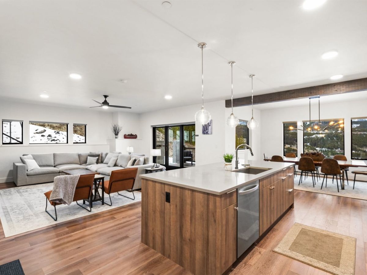 A modern open-concept kitchen and living area with a wood island, pendant lighting, and a spacious gray sectional opens to a dining space with large windows overlooking mountain views.