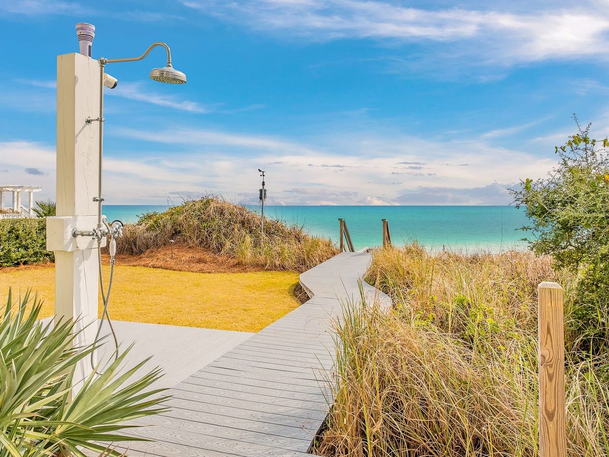 A curved wooden boardwalk leads through tall beach grass to clear turquoise water. An outdoor shower stands nearby, giving the space a relaxed beach-day feel. The view feels bright, peaceful, and perfect for a slow walk to the shore.