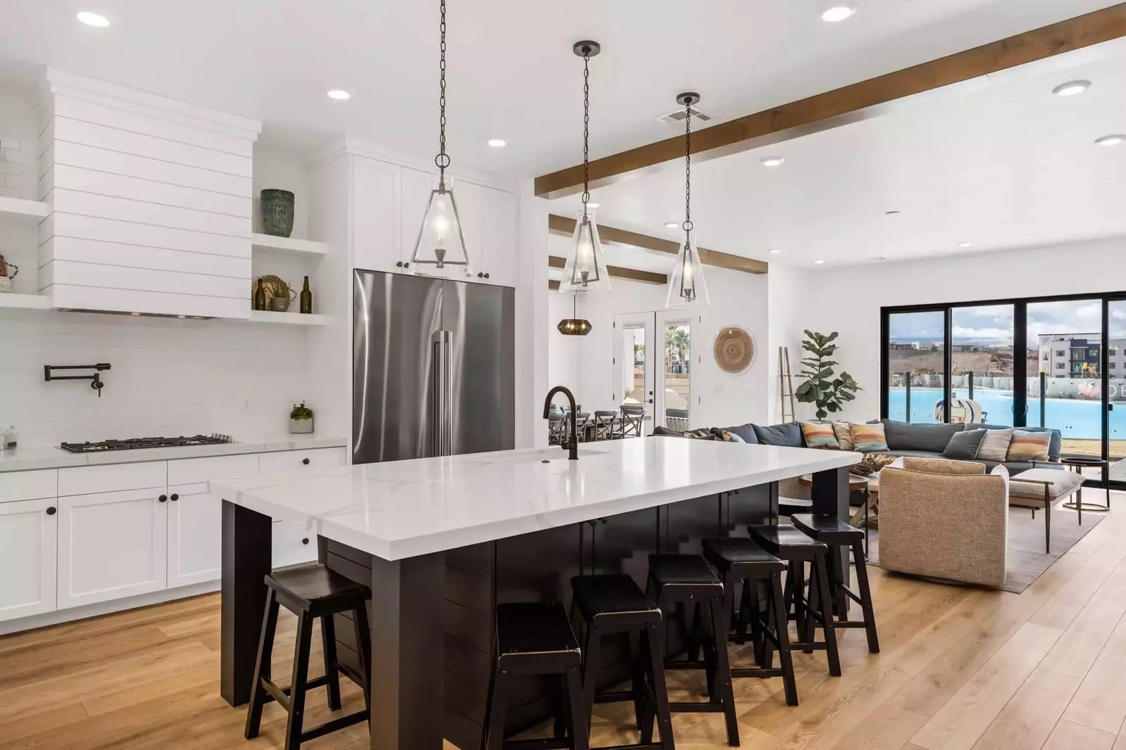 Bright modern kitchen with a large island, black stools, stainless steel appliances, and open living space overlooking a pool.
