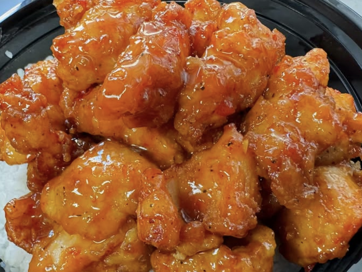 Close-up of crispy orange chicken pieces coated in a glossy sauce, served over a bed of white rice in a black bowl.