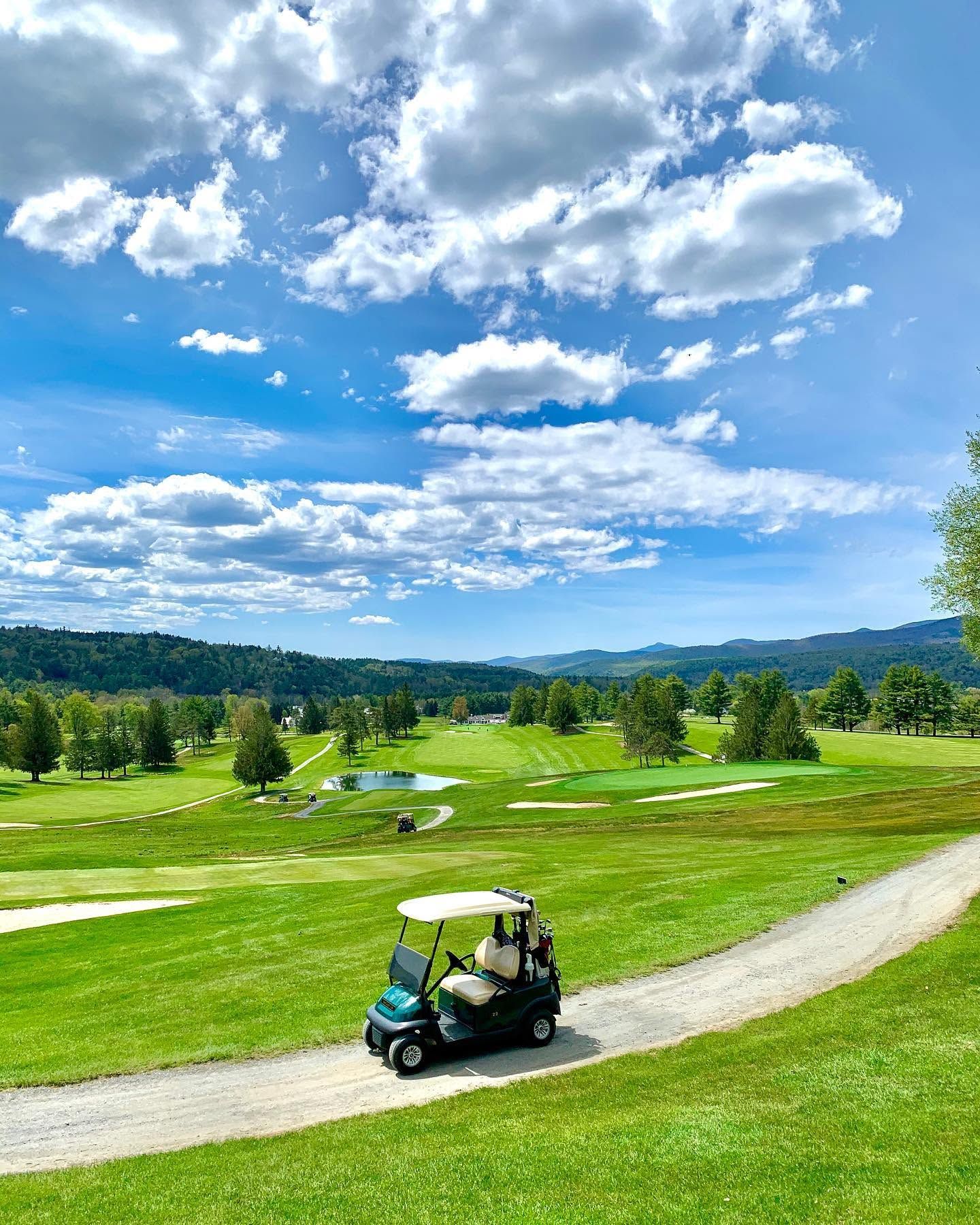 A green golf cart sits on the cart path at Stowe Country Club in Vermont, with a sweeping panoramic view of tree-lined fairways, a sparkling pond, and the Green Mountains stretching across the horizon under a dramatic blue sky filled with billowing clouds.