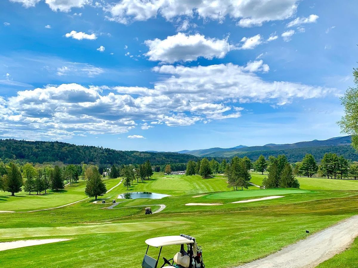 A green golf cart sits on the cart path at Stowe Country Club in Vermont, with a sweeping panoramic view of tree-lined fairways, a sparkling pond, and the Green Mountains stretching across the horizon under a dramatic blue sky filled with billowing clouds.