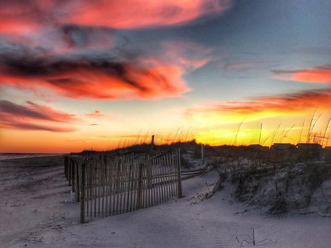 A breathtaking sunset at Holden Beach with fiery pink and orange clouds glowing above the sand dunes and wooden fence. The calm shoreline and golden horizon create a peaceful coastal view.