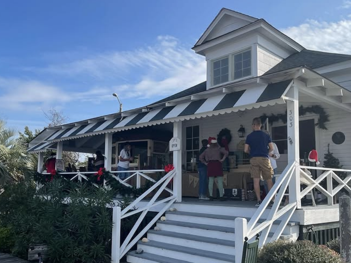 A charming white cottage-style shop with a black-and-white striped awning and festive holiday decorations, including wreaths and garlands, with people gathered on the porch under a bright blue sky.