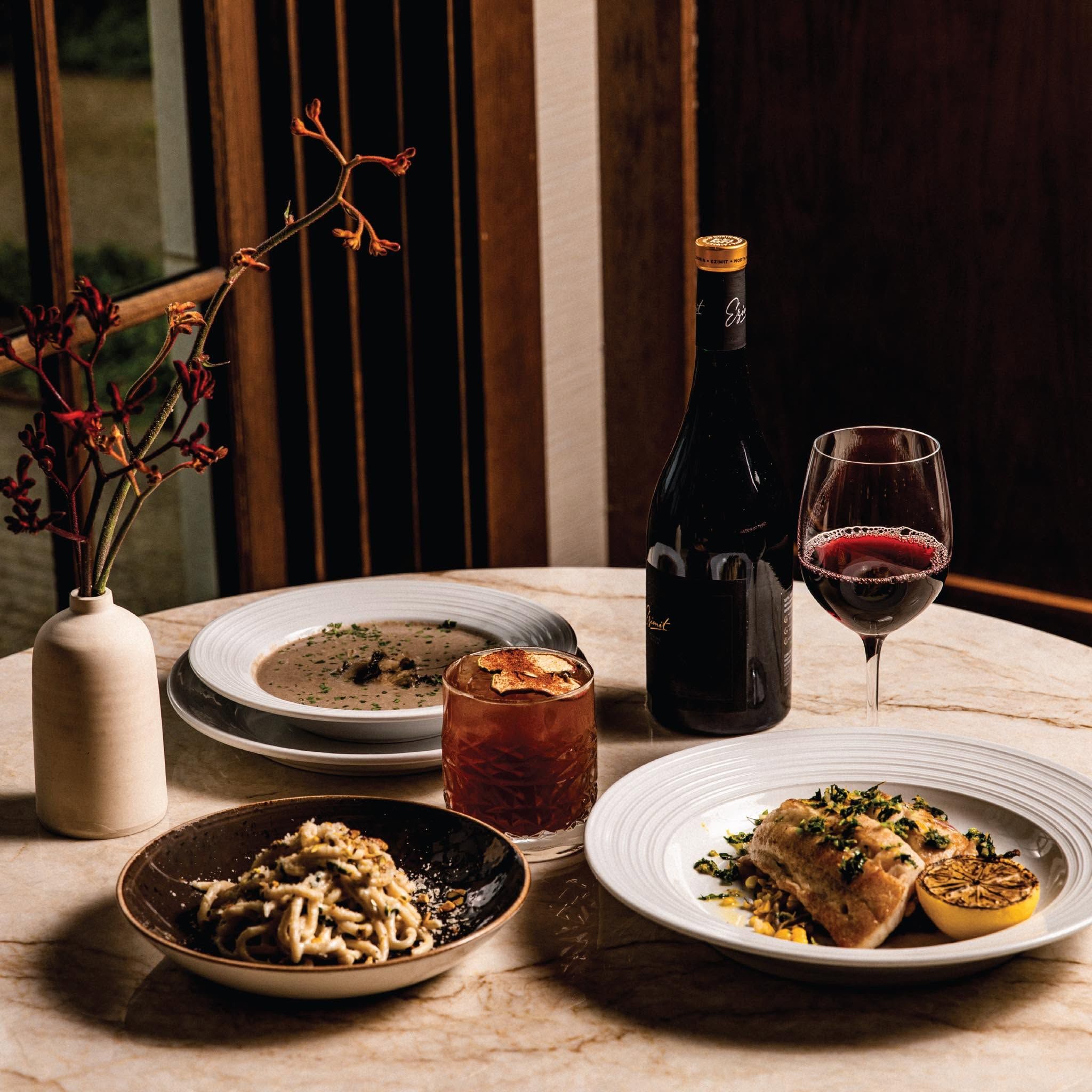 An elegant dinner spread rests on a marble table, featuring creamy pasta, a seared fish fillet with charred lemon, a bowl of mushroom soup, and a craft cocktail beside a bottle and glass of red wine. Warm, moody lighting and a delicate floral arrangement create a refined, intimate dining atmosphere. 