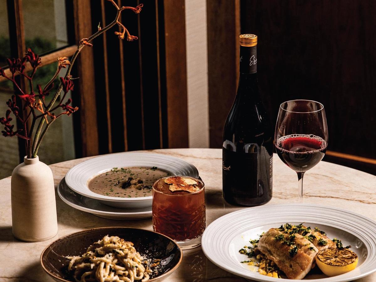 An elegant dinner spread rests on a marble table, featuring creamy pasta, a seared fish fillet with charred lemon, a bowl of mushroom soup, and a craft cocktail beside a bottle and glass of red wine. Warm, moody lighting and a delicate floral arrangement create a refined, intimate dining atmosphere.