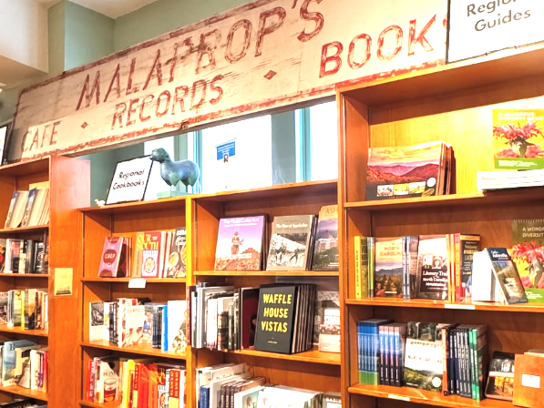 Bookshelves filled with regional cookbooks and travel guides inside Malaprop’s Bookstore, with a rustic sign overhead.