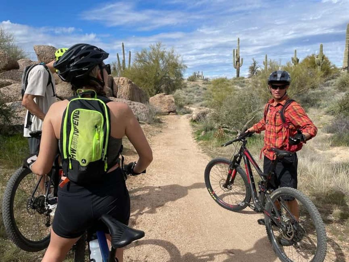 A small group of mountain bikers pause on a desert trail surrounded by towering cacti and rocky terrain. The bright blue sky and open landscape make for an epic outdoor adventure.