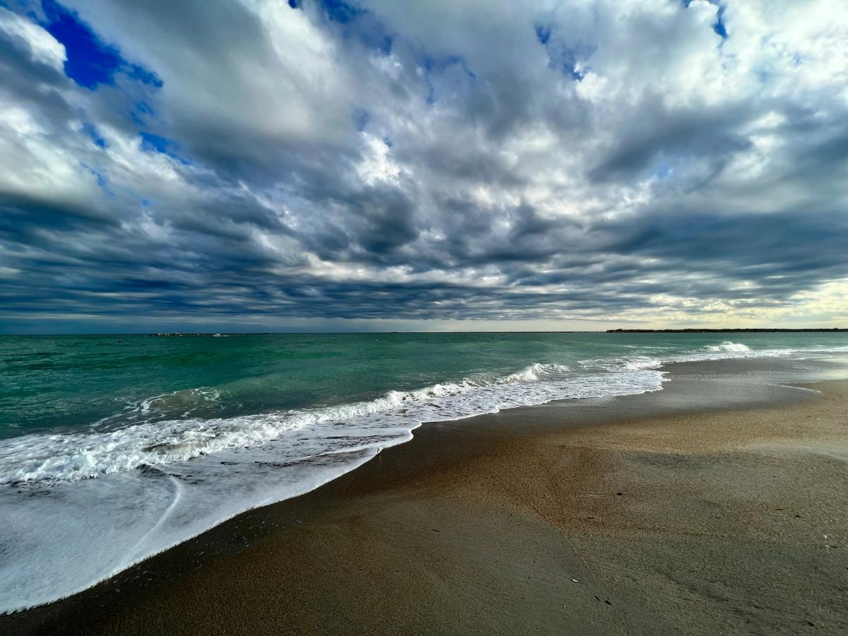 A moody shoreline under dramatic, fast-moving clouds, with turquoise waves gently washing onto a sandy beach.