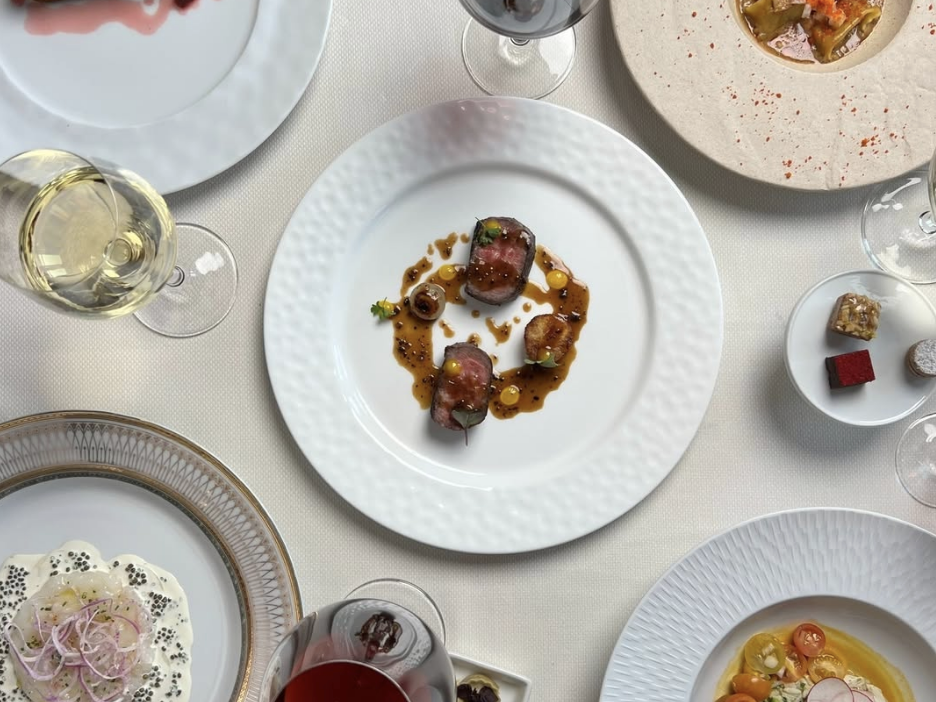 A gourmet dining table set with elegantly plated dishes, including seared meat with sauce, fresh seafood, delicate desserts, and a variety of wine pairings. The table features a white tablecloth, fine china, and artistic plating with vibrant ingredients