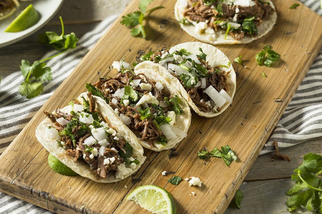 hree soft corn tortilla tacos filled with tender shredded beef barbacoa, diced onions, fresh cilantro, and crumbled cotija cheese line a rustic wooden cutting board. Fresh lime wedges and scattered herbs complete the vibrant Mexican presentation.