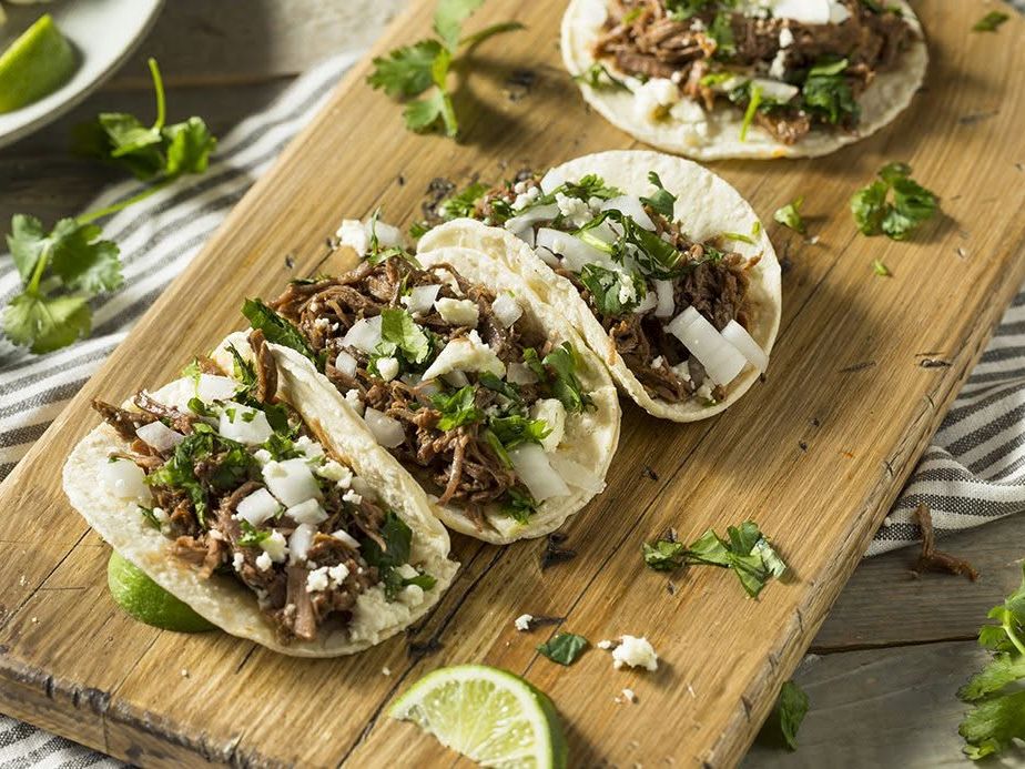 hree soft corn tortilla tacos filled with tender shredded beef barbacoa, diced onions, fresh cilantro, and crumbled cotija cheese line a rustic wooden cutting board. Fresh lime wedges and scattered herbs complete the vibrant Mexican presentation.