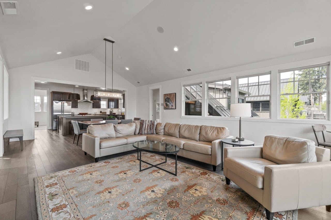 A bright open-concept living room features a beige leather sectional and armchair arranged around a round glass coffee table on a floral area rug, with a vaulted ceiling and a linear chandelier over the adjoining kitchen and dining space. Stainless appliances and a dark wood island complete the layout, highlighting the spacious comfort of this mountain-town vacation rental.