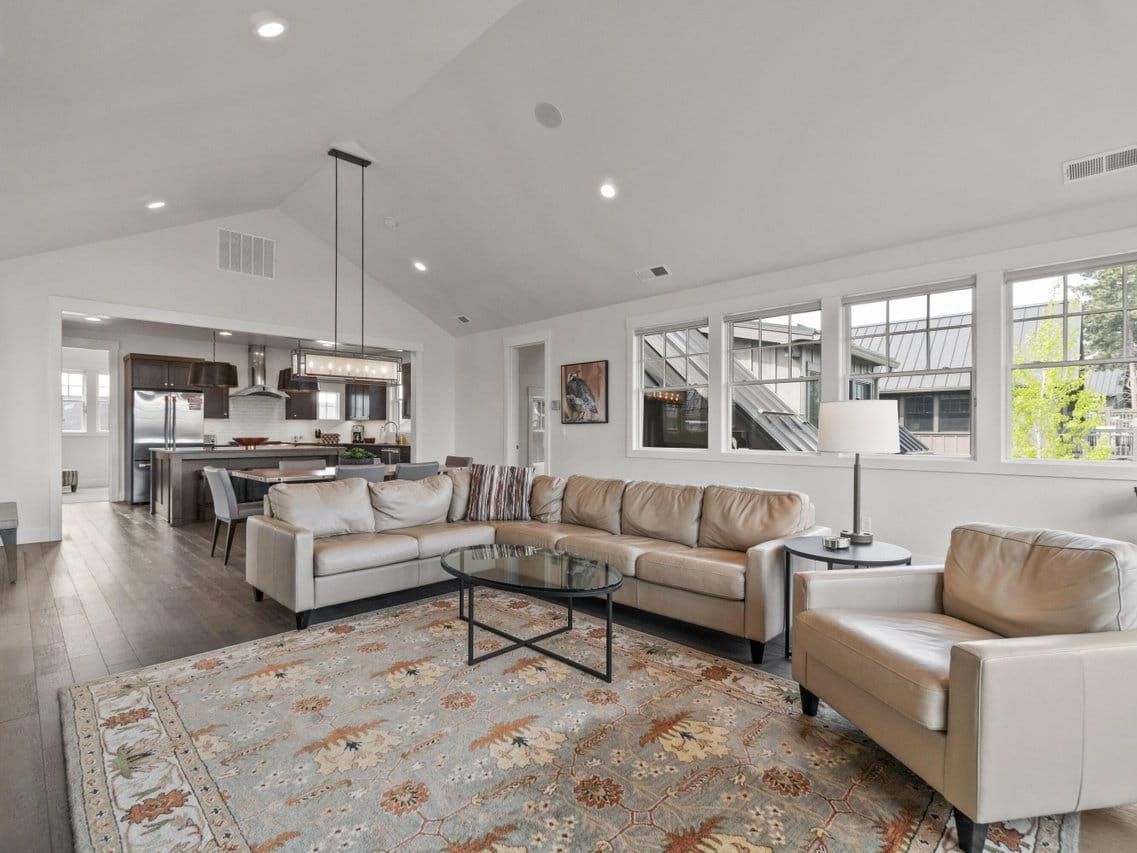 A bright open-concept living room features a beige leather sectional and armchair arranged around a round glass coffee table on a floral area rug, with a vaulted ceiling and a linear chandelier over the adjoining kitchen and dining space. Stainless appliances and a dark wood island complete the layout, highlighting the spacious comfort of this mountain-town vacation rental.