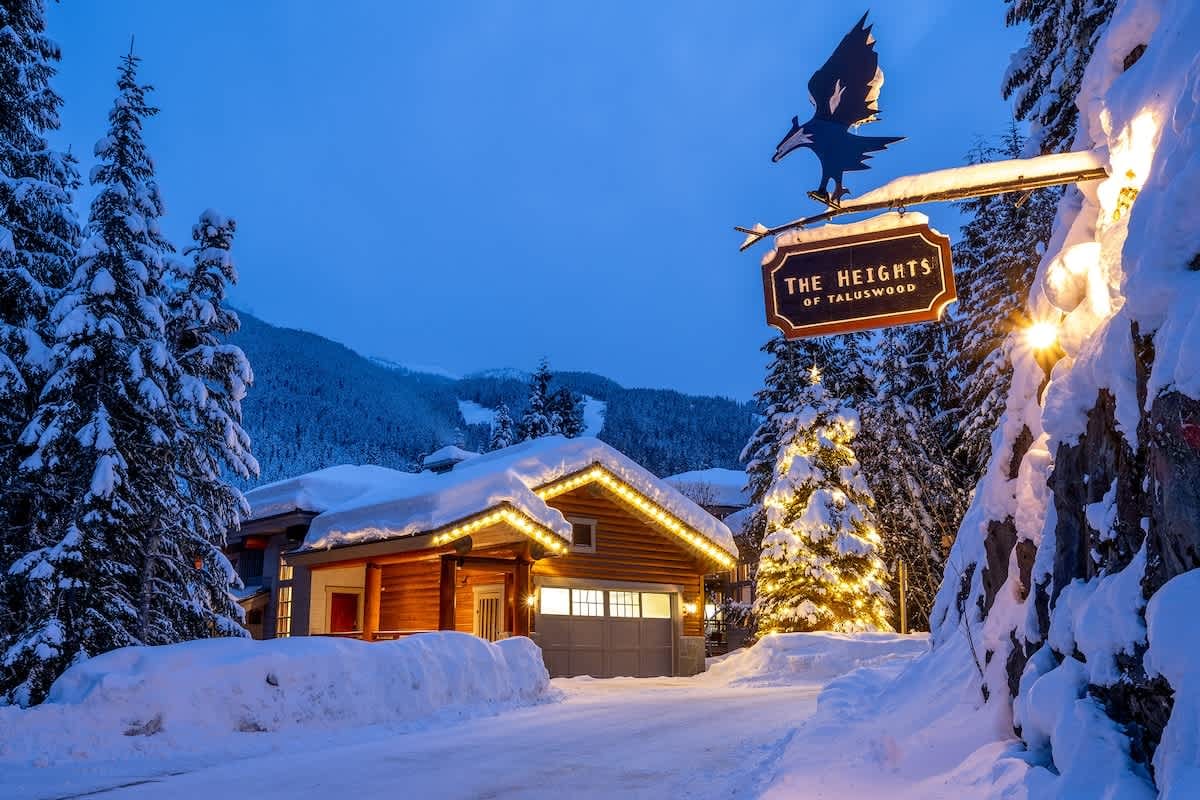 Snow-covered mountain cabin at dusk adorned with warm string lights, framed by towering pine trees and a welcoming illuminated sign in a peaceful winter setting.