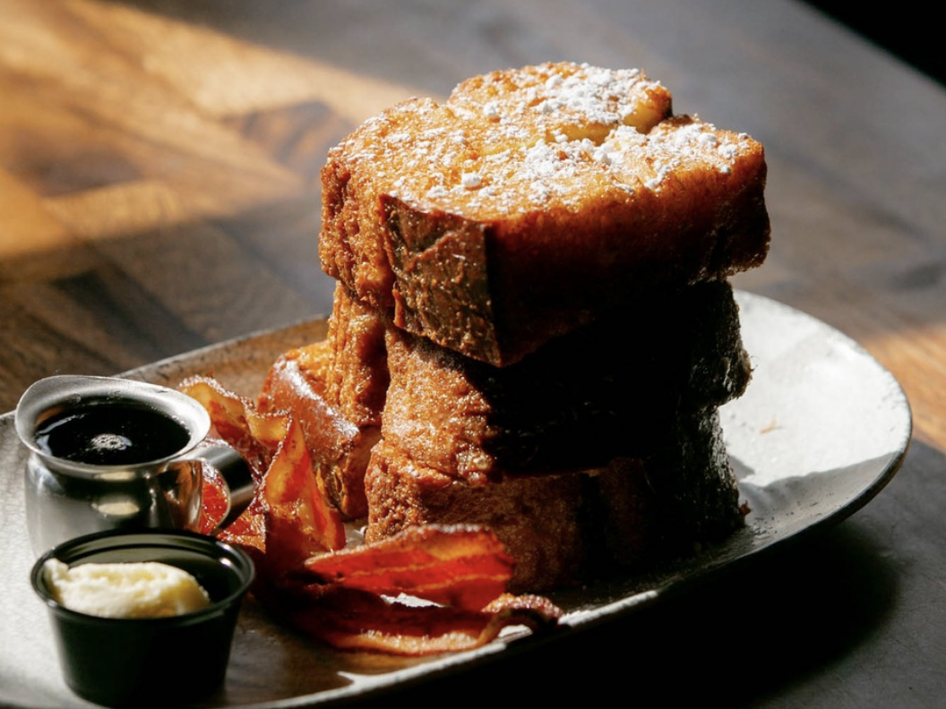 A stack of thick-cut French toast dusted with powdered sugar, served with crispy bacon, syrup, and butter on a rustic plate.