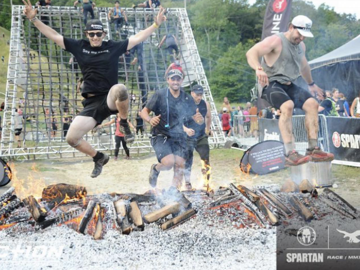 Three men leap over a flaming pit of logs during a Spartan Race, with other participants and a rope wall obstacle visible in the background.
